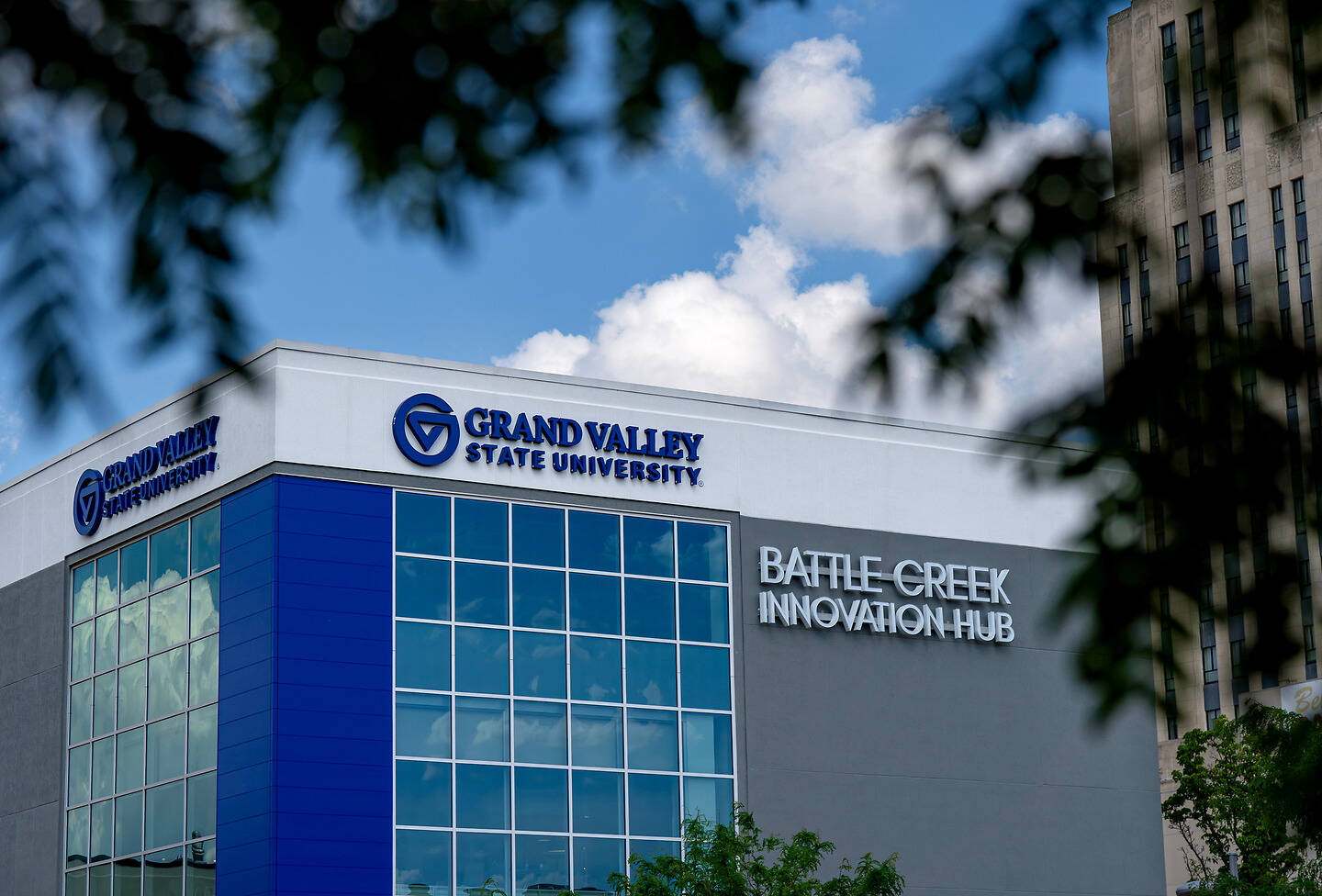 The Grand Valley State University Battle Creek Innovation Hub building is framed by tree branches against a blue sky with clouds.
